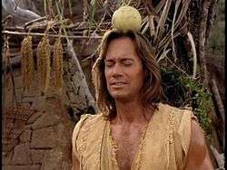 A man with long brown hair stands in a village scene with a piece of fruit on his head, his eyes closed in anticipation.