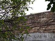 The bridge from the road, looking up through foliage at the east parapet wall with its unornamented date stone.