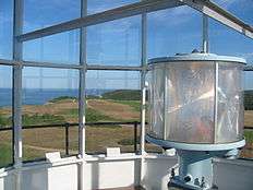 Photograph taken from the lantern room of the lighthouse.  In the foreground on the right is the circular lens surrounding the lamp. The background is the view looking out over the lighthouse grounds and the cliff out to the ocean.