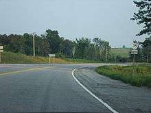 A low perspective of a highway. The highway curves to the right, with rolling hills in the background. A reassurance marker is posted in the ground to the right of the highway