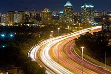 Bird's-eye view of a highway at night. The highway starts at the bottom centre, turning to the right as it progresses into the background. Streams of light show the movement of cars along the highway. Tall poles support lit bulbs. Many buildings and lights are visible in the distance.