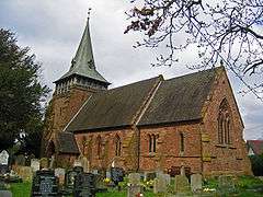 A red sandstone church seen from the southeast. The chancel is slightly lower than the nave and the tower has a broach spire.  Gravestones are in the foreground and part of a tree is to the left