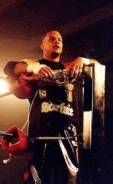 A Hispanic wrestler wearing ring attire: a black graphic shirt with black graphic shorts, taped hands and fingers, and black elbow pads