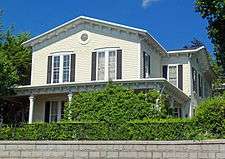 A two-story pale yellow house with black shutters and a slightly pointed roof with brackets along its sides