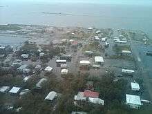 Aerial view of a flooded town. Nearly all roads are flooded, and only buildings, trees, and elevated areas of land can be seen above water. In the background is an ocean.