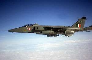 Top forward view of dark-green and blue-grey aircraft overflying sea, above scattered clouds.