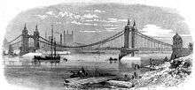 Suspension bridge crossing a wide river. A few buildings can be seen behind the bridge.