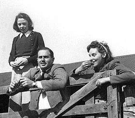 Informal outdoor portrait of dark-haired moustachioed man in suit leaning on fence, flanked by two dark-haired women