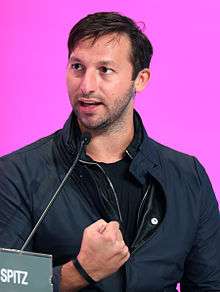 Young man with short blonde-brown hair wearing a dark open necked shirt and zip-up jacket, not cleanly shaven, smiling broadly. The background is labelled with the word 'Brasserie'.