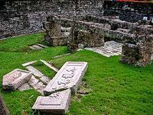 On a green lawn surrounded by a brick wall lie broken engraved stones and stone foundations.