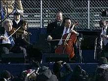 Four male musicians playing a variety of instruments outdoors in front of a chain link fence