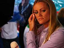 Woman with long dark eyelashes, long brown/blonde hair wearing a lavender shirt and with hand on hair, and large bangle earrings, looks sideways.