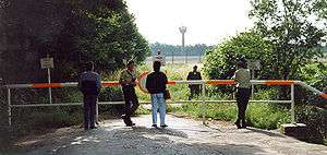 View of a road terminating in a red and white horizontal barrier, with trees on either side. Four people, two in uniform, are standing on the near side of the barrier. On the far side is another uniformed man standing in a grassy field. In the far background is a high metal fence and a tall watchtower with an octagonal cabin at its top.