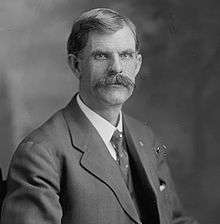 A man with dark hair and a prominent mustache wearing a black jacket, patterned tie, and white shirt