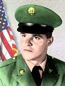 Head and shoulders of a young white man looking off to the side, wearing a peaked cap and a military jacket over a shirt and tie.