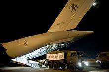 Colour photo of a truck with a sign marked with Japanese characters over its cargo compartment driving away from the rear ramp of a grey aircraft at night