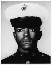 Head of a young black man wearing a dark military jacket and a white peaked cap with a black visor.