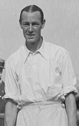 A black-haired man in a white shirt with sleeves rolled up looks into the camera, posing