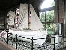  White-hulled small boat sitting on a base of stones behind a rail, within a museum. The name "James Caird" is visible. A stuffed penguin in a glass case stands nearby.