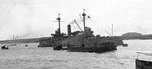 Black and white photo of a warship with a small amount of smoke coming out of its funnel. Several smaller ships are moored near the warship, and land is visible in the background.