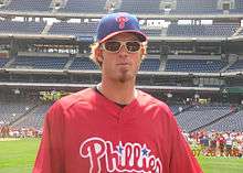 A man with an extended soul patch wearing a red baseball jersey and a blue baseball cap with red brim and a red "P" on the face