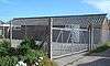 Side view of a long, low brick building with a corrugated roof, behind a wire fence and metal gates and next to an asphalt car park.  There are two doors and two sets of square windows.