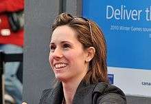 Young smiling brunette with shoulder-length hair held back by a pair of sunglasses on top of her head. She is wearing a dark gray coat and behind her is a blue-and-white outdoor ad about the 2010 Winter Games.