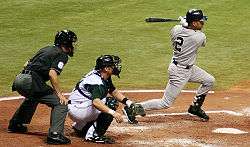 A baseball player in a grey uniform follows through with his swing, while a catcher on the opposing team in a white uniform squats behind him and an umpire in a black uniform stands behind the catcher.