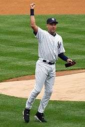 A man in a white baseball uniform with navy pinstripes raises his right arm in the air while holding a box in his left hand.