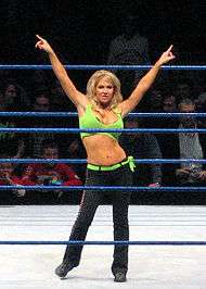 A blond woman poses in a wrestling ring with blue ropes. Her arms are outstretched, and she is pointing to either side. She is wearing a green crop top, with a matching green belt, and black trousers.