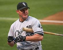 A man wearing a gray baseball uniform with white and black trim and a black baseball cap with "Sox" in white Old English lettering on the face holding a baseball bat under his left arm