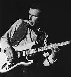 A black-and-white shot of a 36 year-old-man. He is playing an electric guitar at a live concert. His body is tilted to his left, with his face twisted to look down and to the right.