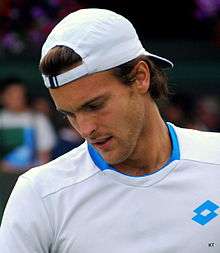 João Sousa in a white shirt and cap looks down before a serve