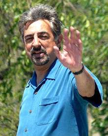 Colored photograph of a man, waving with his left arm. He is wearing a blue shirt.