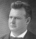 Photographic portrait of a man about forty, with short hair standing straight. He is dressed formally with a bow tie visible, and is facing somewhat to the left.