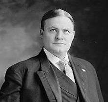 A man with thin, black hair wearing a black jacket and vest, patterned tie, and white shirt