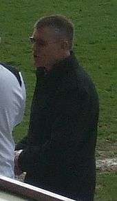 A man with grey hair is wearing a black jacket. He is standing on a grass field.