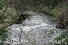 A swiftly-flowing stream about 15 feet (4.5 meters) wide goes around a bend between raised banks. A low retaining wall protects the outer bank.