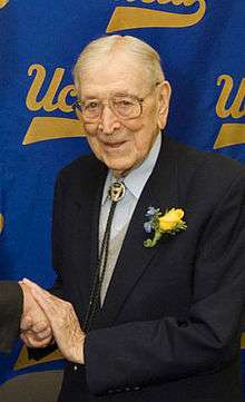  A smiling, elderly man is shown from the waist up. He is shaking someone's hand, but that person is out of the picture. The man is wearing a dark suit with a yellow boutonniere. He has thin white hair and large glasses. He is standing in front of a blue screen that has the script "UCLA" logo on it in yellow letters.