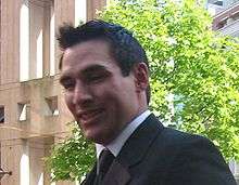 Chest view of a man with short black hair and wearing a suit. He smiles as he looks at the photographer.