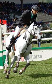 A man riding a white horse at a canter after the horse has crossed a hurdle