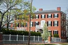 A three story brick house with white trim and black shutters. The front door is sheltered by a portico, which also supports a second floor balcony.