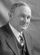 A man with thinning, salt-and-pepper hair wearing a black jacket and vest, white shirt, and patterned tie