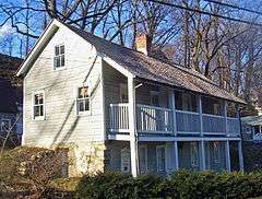 A two-story light green wooden building with a full-length front balcony and exposed stone foundation, seen from its right corner.