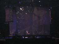 A stage with a big screen display showing a man with a beard. Sitting in a piano, a man with long curly hair wears a white shirt, with the sleeves roll back, and a black vest, plays the piano. To the man's left, a man can be seen standing playing an electric piano.