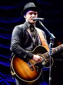 A blonde man is wearing a suit and playing a guitar while singing on a microphone