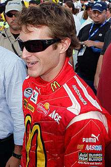 A man in his late twenties wearing black sunglasses with a head full of hair and a red jacket with sponsors' logos.