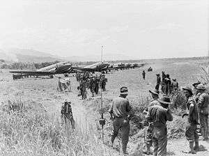 A line of Dakotas on a grass airstrip. Men wearing slouch hats file off a plane. A jeep drives along the strip. Other men in uniform and civilians look on.