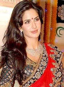 A woman dressed in traditional Indian clothing is looking away from the camera. She has a red bindi between her eyebrows, a silver necklace and is wearing an embroidered red sari.