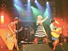 Four people on a stage; three are standing at the front of the stage behind microphone stands. At the back of the stage, one woman is sitting behind a drum set.